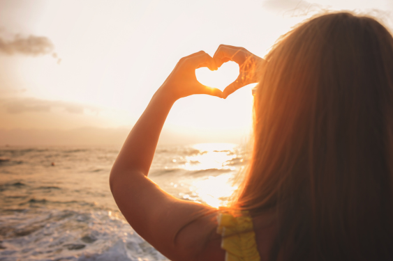 A woman makes a heart gesture to the sun over the sea showcasing the importance of self-love